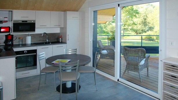 A modern kitchen with built-in appliances, a dining area with a round table and chairs, and a sliding glass door leading to an outdoor patio with wicker furniture. - LE Chalet - Bazeilles (BAZEILLES)