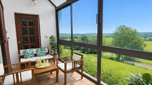 A conservatory with a view of a green landscape, a wooden table with chairs, and a window with a view of the countryside.