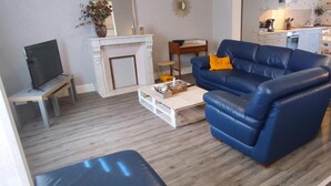 Ein Wohnzimmer mit blauem Sofa, weißem Couchtisch, Fernseher auf einem Schrank, Kamin und einer Küche im Hintergrund.