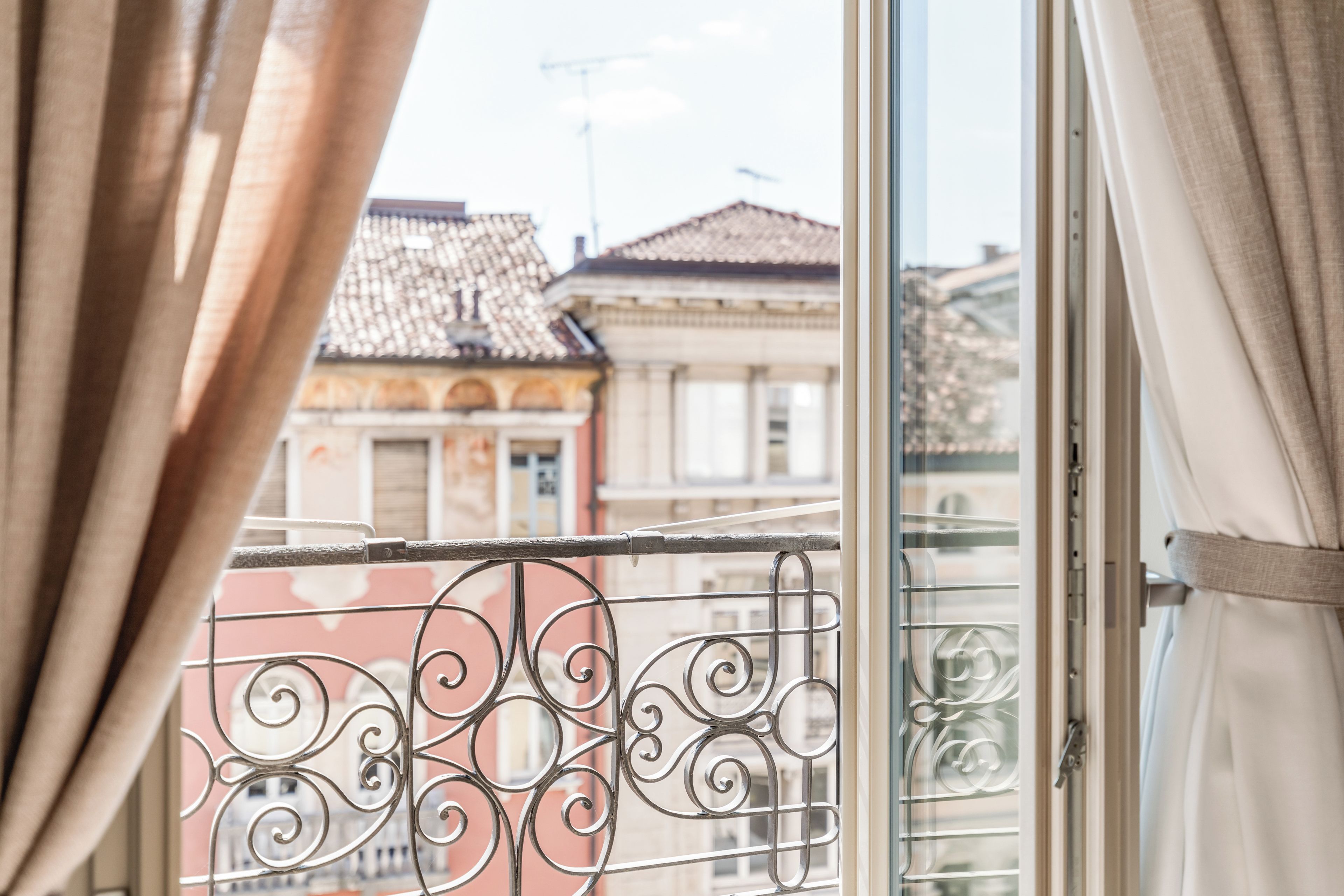 A window with metal railings and curtains, offering a view of buildings with traditional architecture.