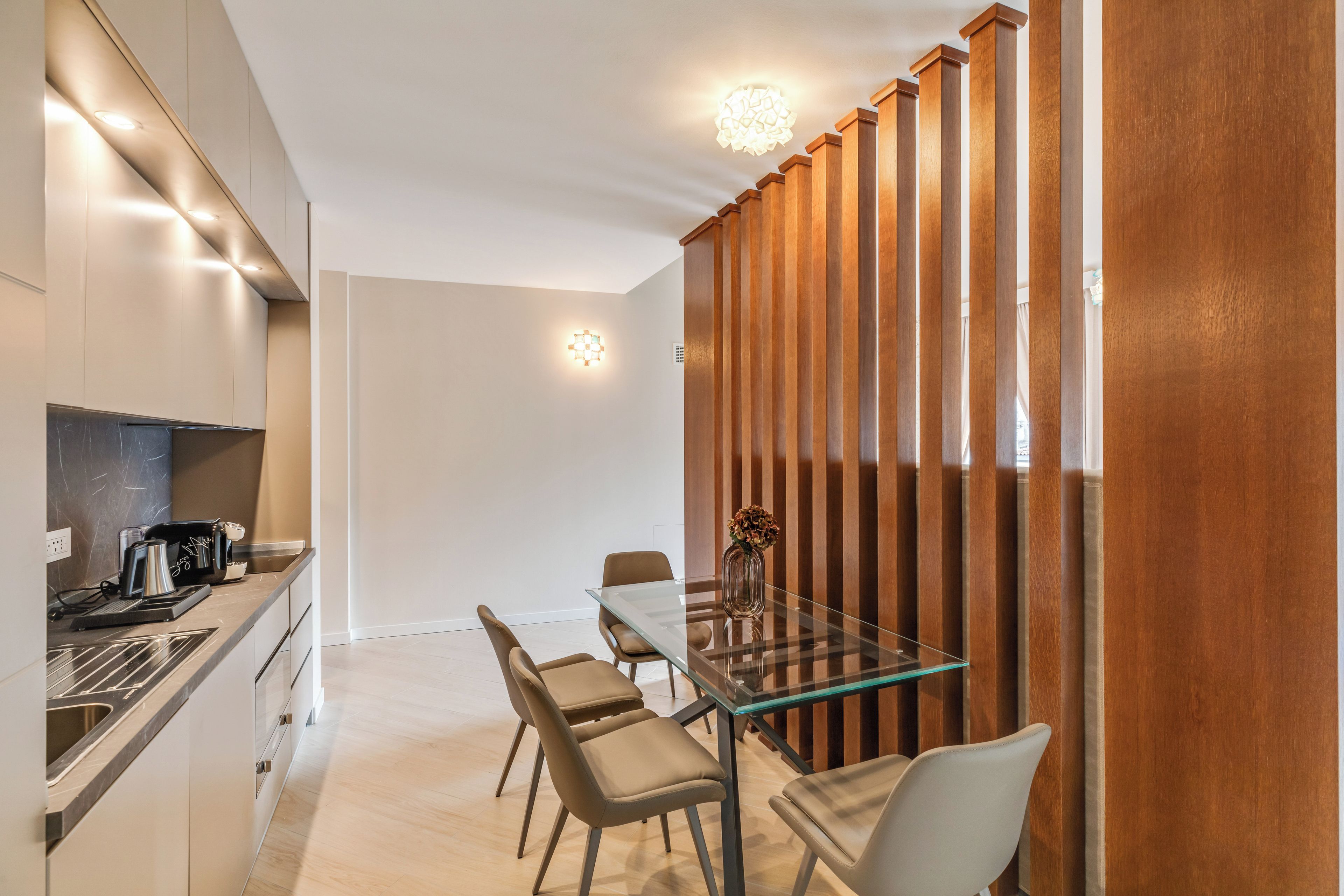 Una cocina moderna con estufa integrada, cafetera y pileta. Hay un comedor con mesa de vidrio y cuatro sillas.