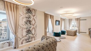 Una sala de estar moderna con un amplio sofá modular, un otoman tapizado y un sillón verde azulado. La habitación cuenta con un gran ventanal con cortinas, un mural decorativo y una lámpara de techo circular.