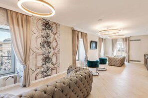 A modern living room with a large sectional sofa, a tufted ottoman, and a teal armchair. The room features a large window with curtains, a decorative wall mural, and a circular ceiling light fixture. - Oriana Homèl Udine (Udine)