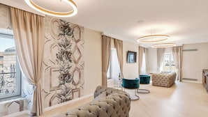 A modern living room with a large sectional sofa, a tufted ottoman, and a teal armchair. The room features a large window with curtains, a decorative wall mural, and a circular ceiling light fixture. - Oriana Homèl Udine (Udine)
