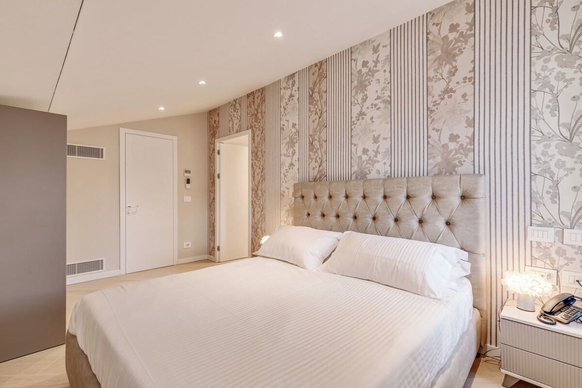 Un dormitorio con una cama grande, un cabecero capitonado, una mesita de noche con lámpara y una puerta con ventana.