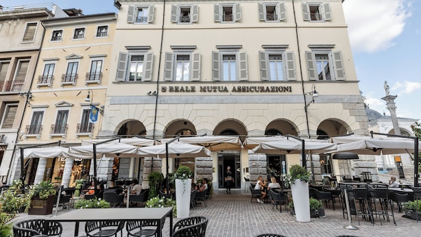 A building with a sign reading "S. Reale Mutua Assicurazioni" and outdoor seating with umbrellas. - Oriana Homèl Udine (Udine)