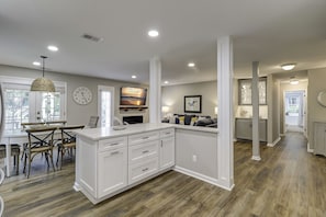 Une cuisine moderne avec des meubles blancs, un coin repas avec une table et des chaises en bois, et un salon avec un canapé et un téléviseur.