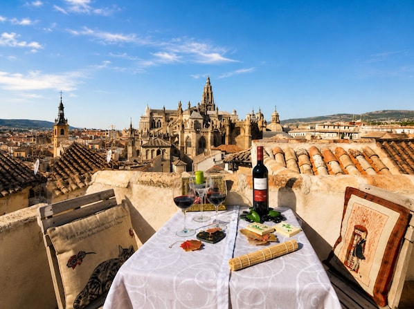 A table set with a bottle of wine, glasses, and food items on a terrace overlooking a historic cityscape with a prominent cathedral.