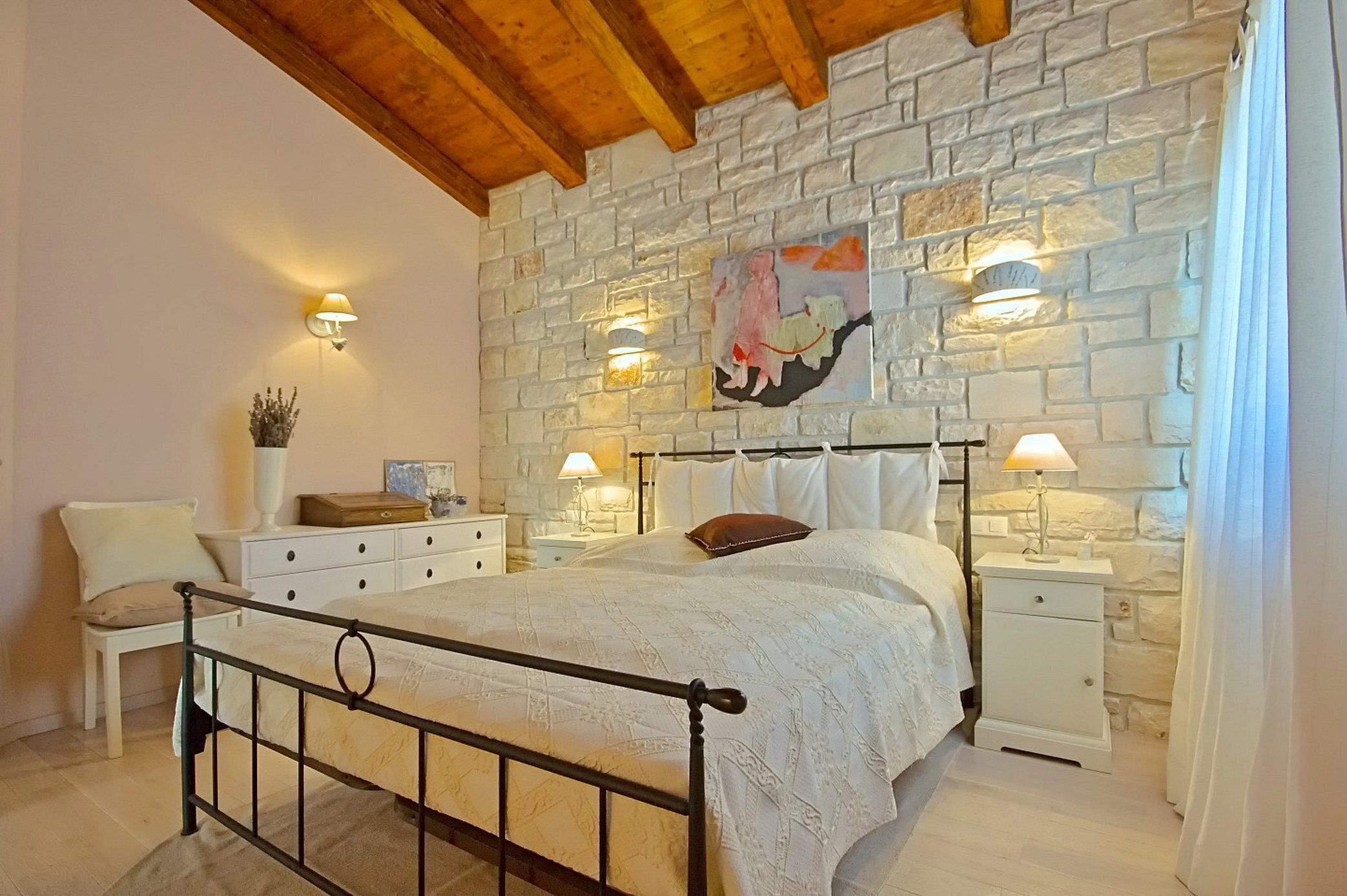 Ein Schlafzimmer mit Steinsturz, einem Bett mit weißen Leinen, zwei Nachttischen mit Lampen, einem Beistelltisch und einem Sessel.