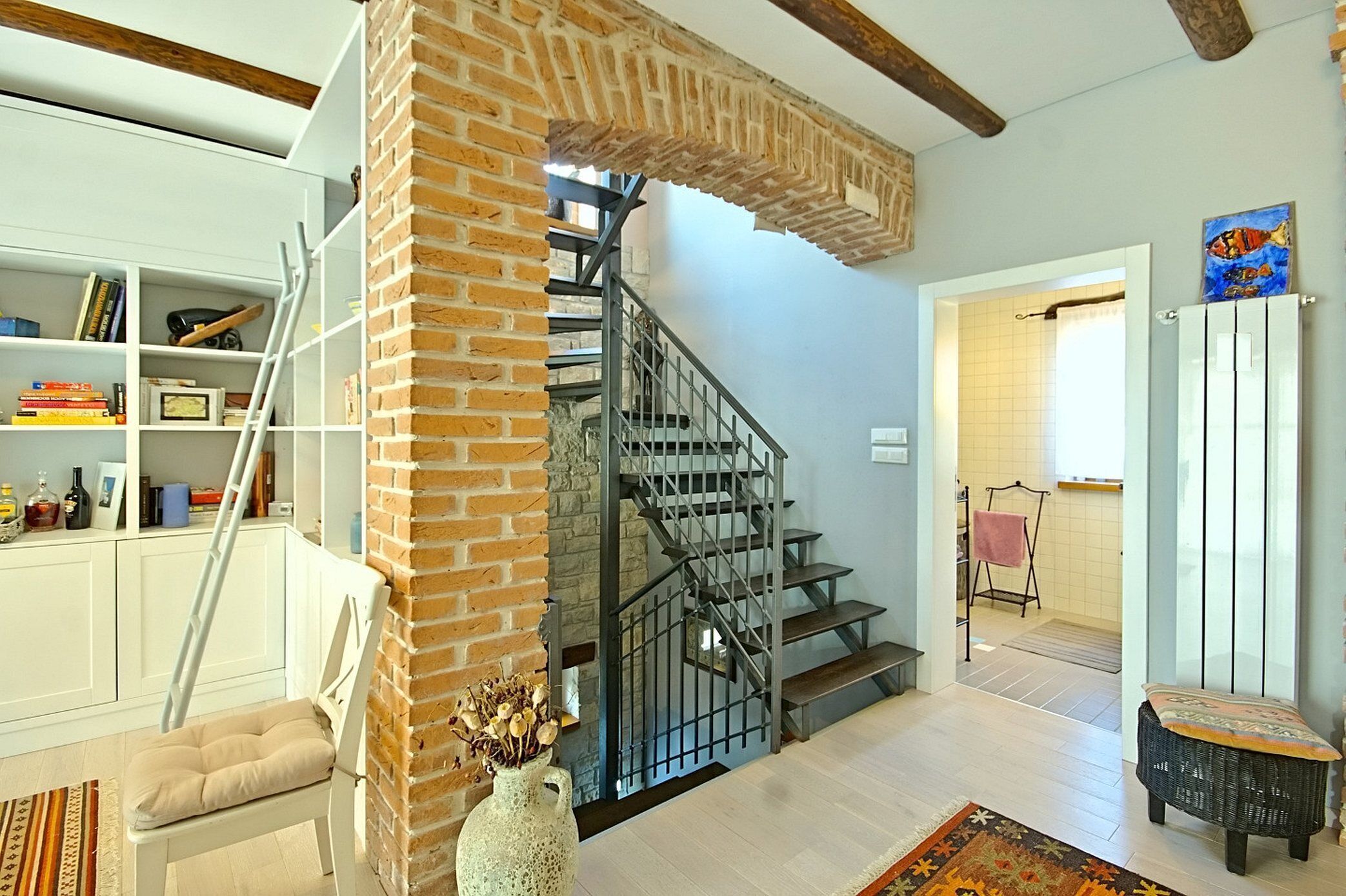 A staircase with a brick wall, a white chair, a bookshelf, and a painting.