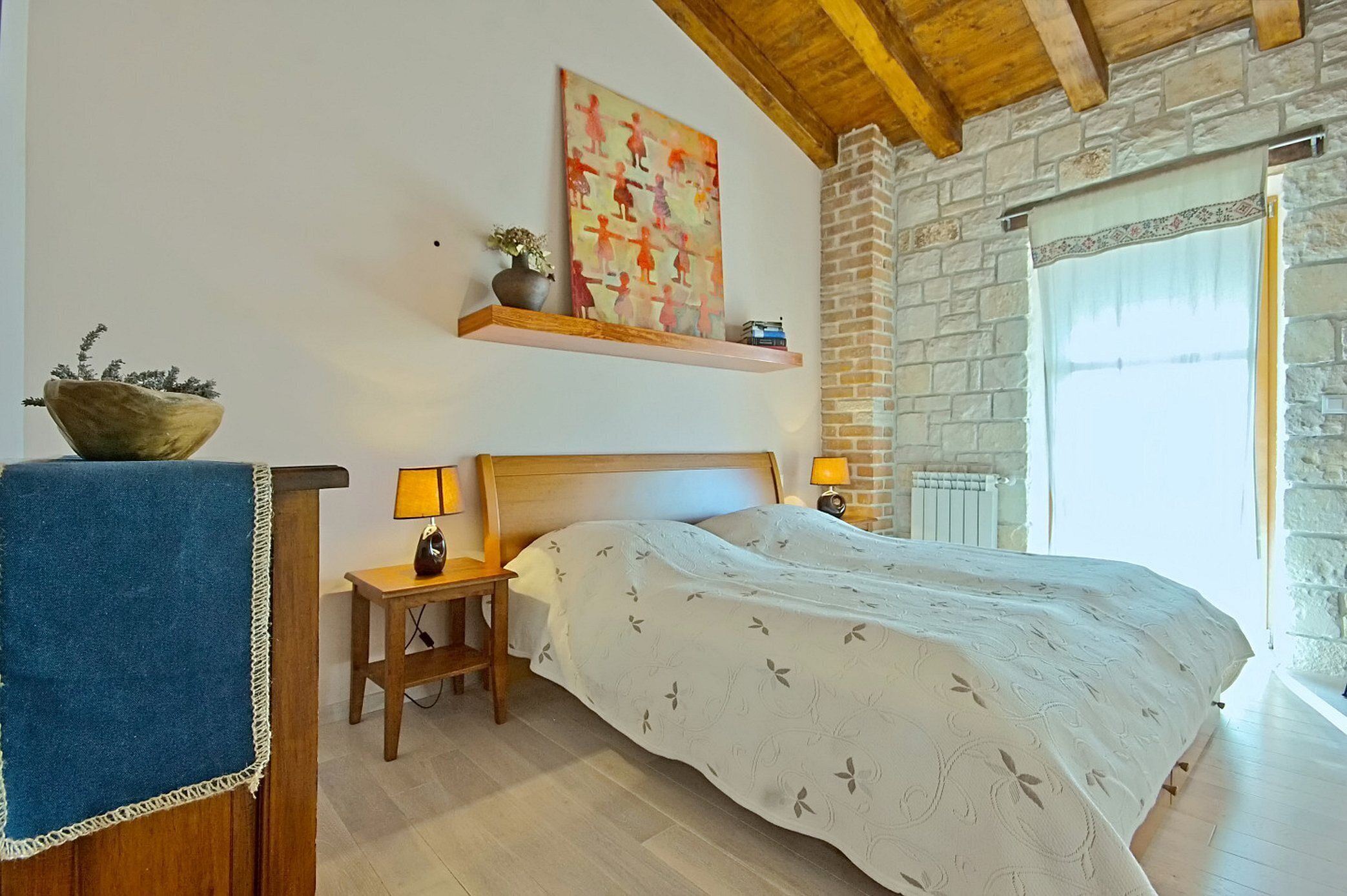 A bedroom with a wooden bed, a nightstand with a lamp, a shelf with a painting, and a stone wall.