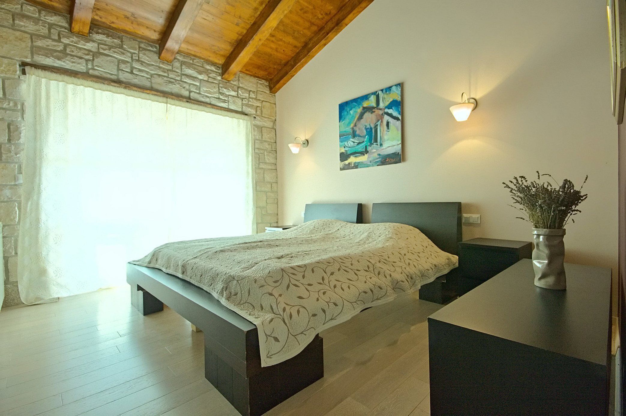A bedroom with a bed, a stone wall, a painting, and a vase with flowers.