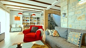 A cozy living room with a red armchair, a grey sofa, and a coffee table with a candle. There is a bookshelf filled with books and decorative items, and a brick wall with a staircase. - Sea view, garden with barbecue, pool with loungers, parking, air conditioning, wifi (Rojci)
