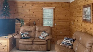 Ein gemütliches Wohnzimmer mit einem braunen Ledersofa, einem kleineren Ledersessel und einem Fernseher auf einem Holzboden.