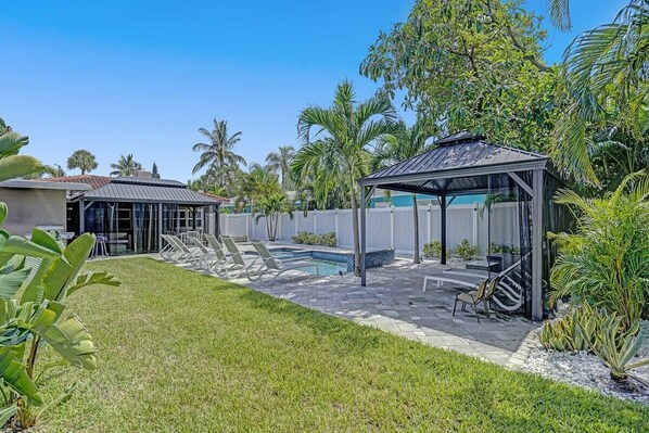 Un jardin avec une piscine, des chaises longues, un kiosque et des plantes tropicales.