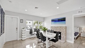 A modern dining area with a white table and chairs, a stone fireplace, and a view of the outdoors through large windows. - Ocean Breeze Villa 🏝️ 5 Minute Walk to Beach (Pompano Beach)