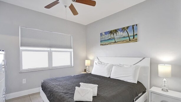 A bedroom with a bed, a ceiling fan, a window with blinds, a painting of a beach, and a nightstand with a lamp. - Ocean Breeze Villa 🏝️ 5 Minute Walk to Beach (Pompano Beach)