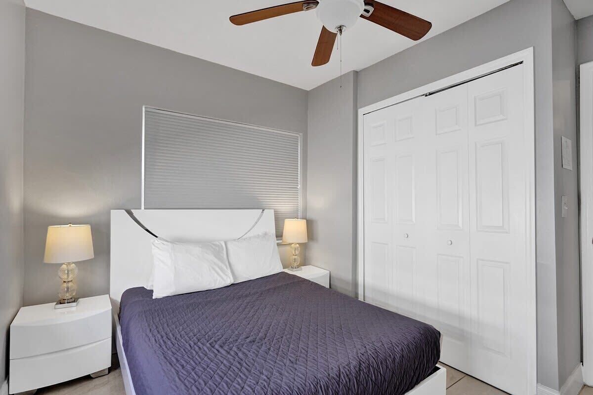 Une chambre à coucher comprenant un lit, deux tables de chevet avec des lampes, un ventilateur de plafond, une fenêtre avec des stores et une armoire.