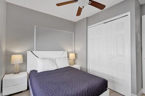 Une chambre à coucher comprenant un lit, deux tables de chevet avec des lampes, un ventilateur de plafond, une fenêtre avec des stores et une armoire.