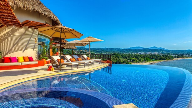 A infinity pool with lounge chairs and umbrellas, a thatched roof structure, and a view of a beach and mountains. - Above Villa Paloma Blanca I Pool/Spa I Beach Club I Views I Spa (LA CRUZ DE HUANACAXTLE)