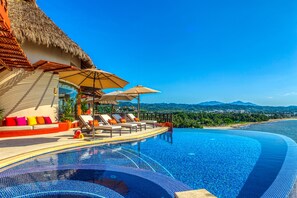 A infinity pool with lounge chairs and umbrellas, a thatched roof structure, and a view of a beach and mountains.