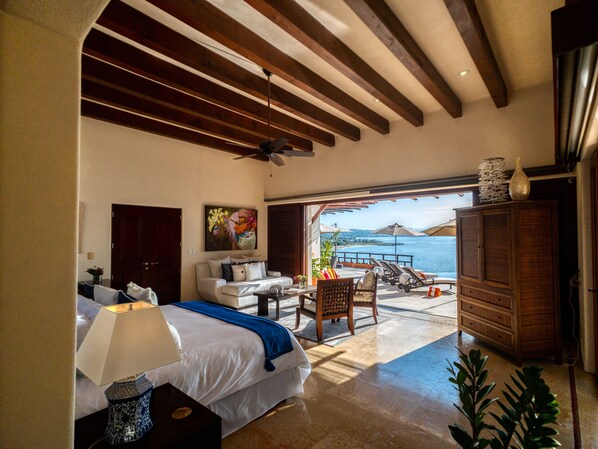 A bedroom with a bed, a desk, a chair, a nightstand, and a view of the sea.
