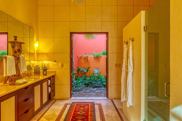 A bathroom with a tiled floor, a sink with a mirror, a towel rack, and a door leading to a room with a red wall and plants.