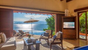 A room with a large window overlooking a body of water, furnished with a sofa, chairs, and a TV. - Above Villa Paloma Blanca I Pool/Spa I Beach Club I Views I Spa (LA CRUZ DE HUANACAXTLE)