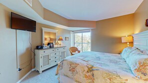 A bedroom with a large bed, a dresser, a television mounted on the wall, and a window providing natural light.