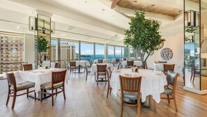 A spacious dining area with round tables and chairs, large windows offering a view of the sea, and a ceiling fan.