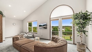 Ein geräumiges Wohnzimmer mit einer großen Couch, einem kleinen runden Tisch und einer Topfpflanze. Zwei Fenster bieten Blick ins Grüne und es gibt einen Balkon.