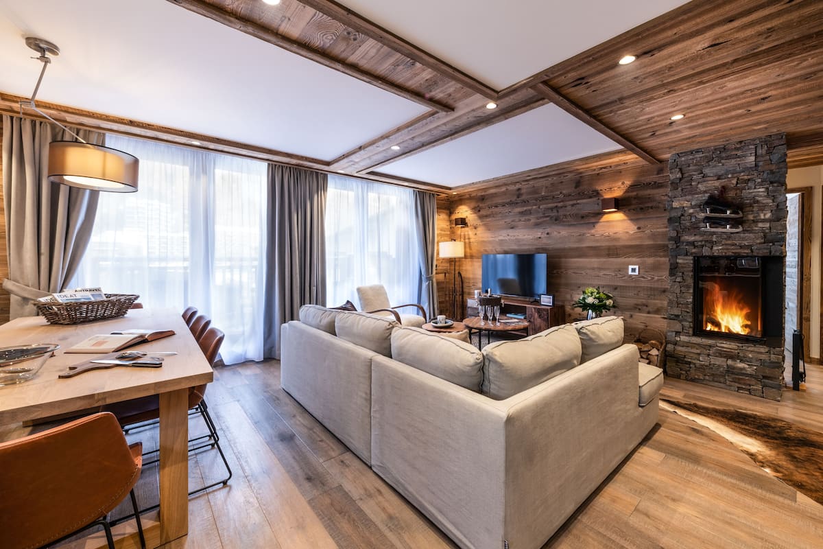 Ein modernes Wohnzimmer mit Kamin aus Stein, einem großen Sofa, einem Esstisch mit Stühlen und einem Fernseher.