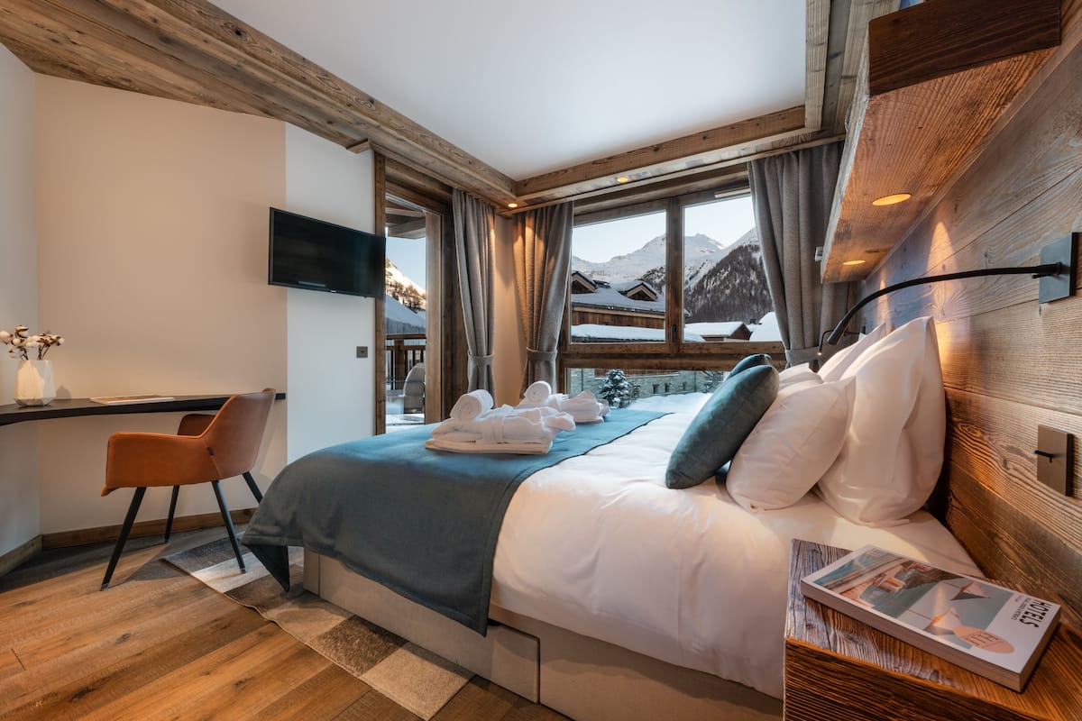 Ein modernes Hotelzimmer mit Holzboden, einem großen Bett mit weißer Bettwäsche, einem Schreibtisch mit Stuhl und einem an der Wand befestigten Fernseher.