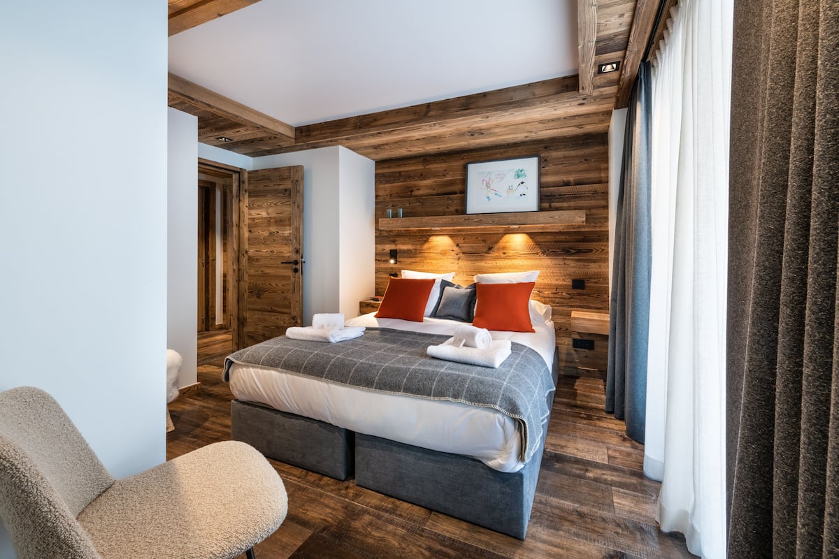 A modern hotel room with a wooden ceiling, a large bed with pillows, a bedside table, and a chair.