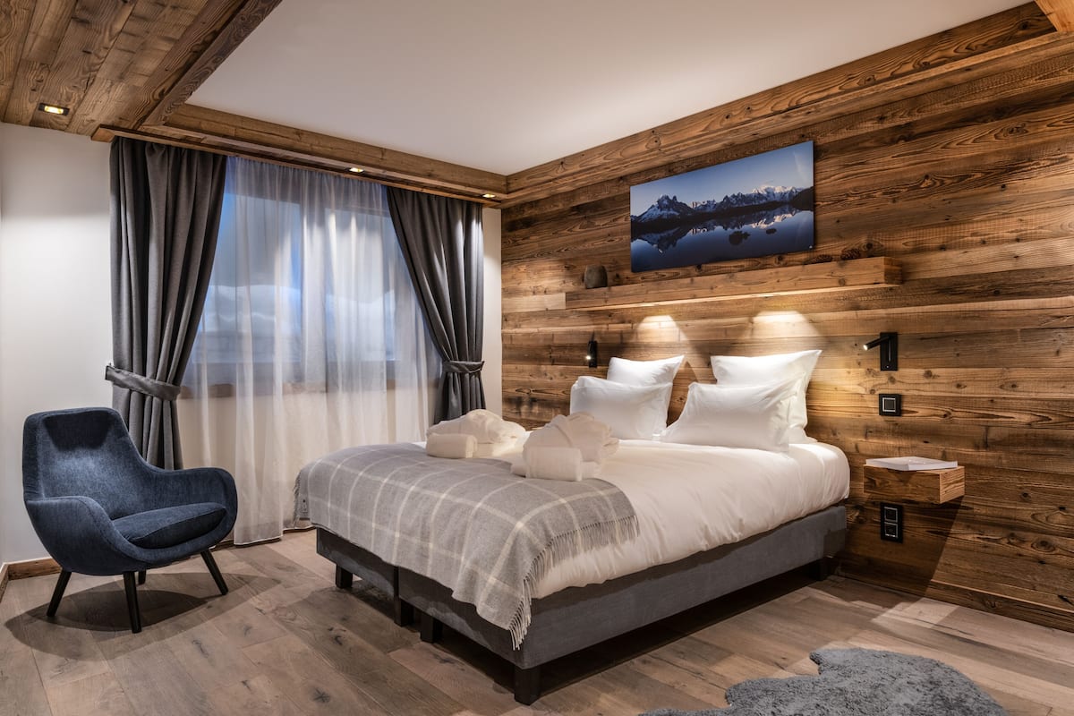 A bedroom with a wooden wall panel, a bed with white bedding, a blue armchair, and a nightstand with a lamp.