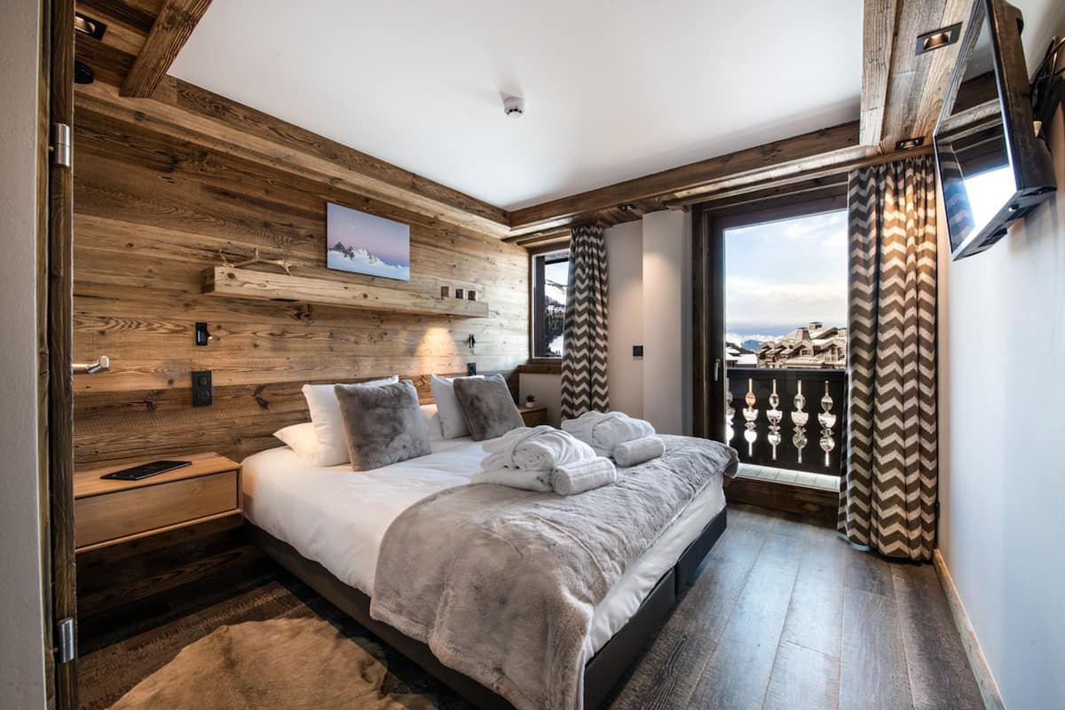 A bedroom with a wooden wall, a large bed, a nightstand, a TV, and a view of the outdoors.
