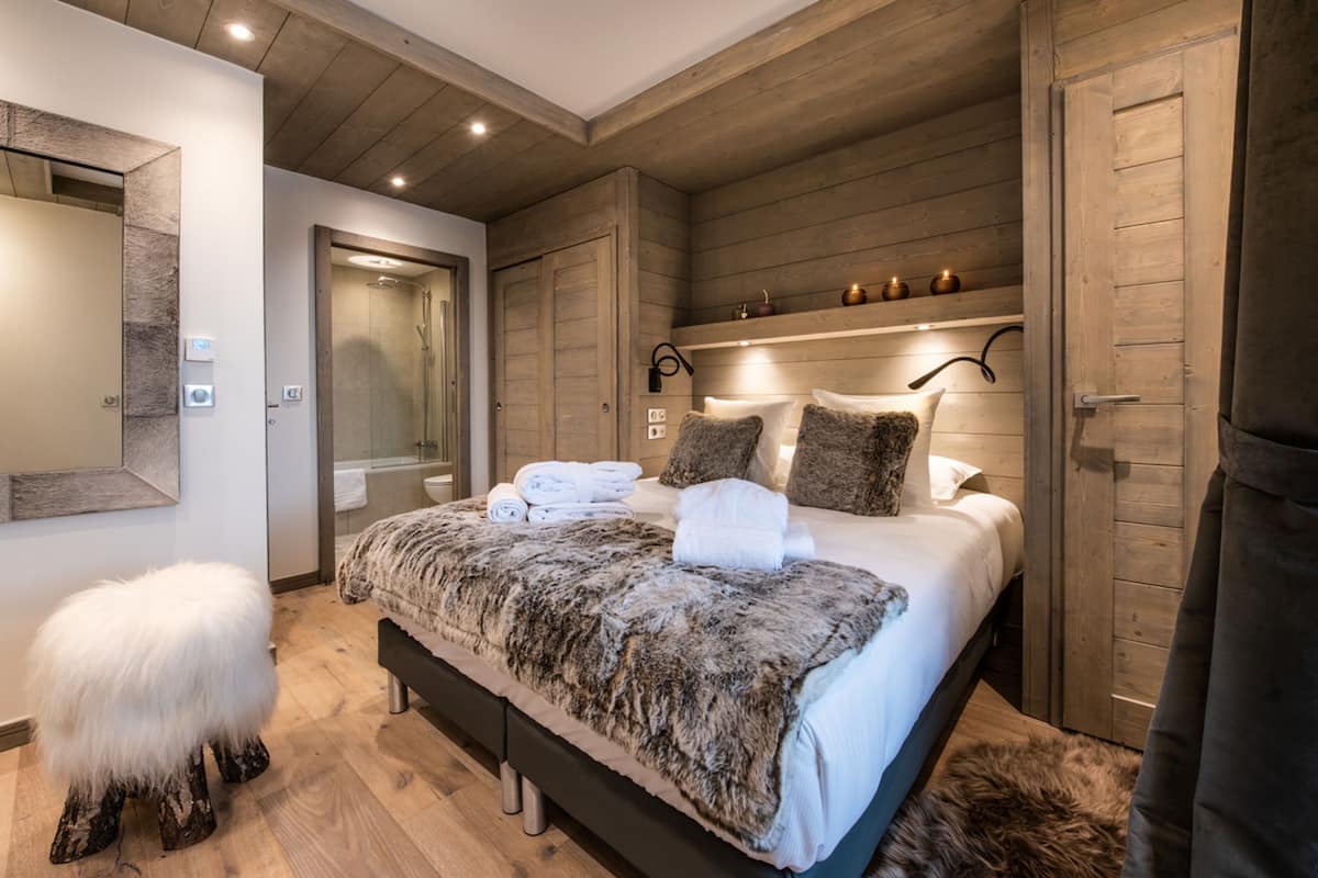 A modern bedroom with a large bed, wooden paneling, a small stool, and a shower area.