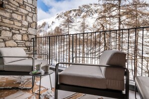 A balcony with a stone wall, a cushioned chair, a small table with a mug, and a view of snow-covered trees and mountains. - Vail Lodge by Alpine Resorts (Val-d'Isere)