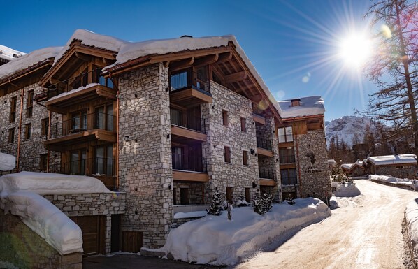 A multi-story stone building with balconies, a ski slope in the background, and snow-covered surroundings. - Vail Lodge by Alpine Resorts (Val-d'Isere)