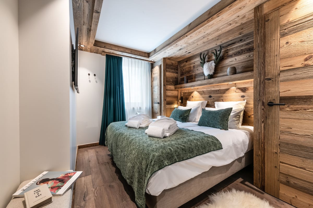 A bedroom with a wooden ceiling, a bed with green bedding, a white door, and a shelf with decorative items.