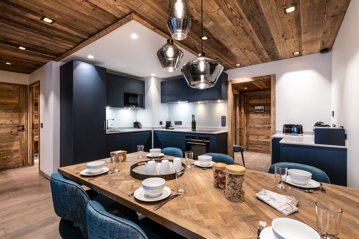 A modern kitchen with wooden ceiling, dark blue cabinets, and a dining area with a wooden table set for four.