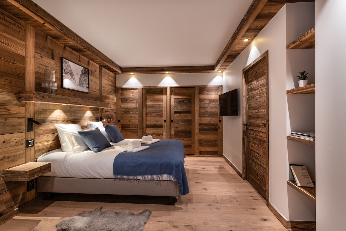 A bedroom with a wooden ceiling, a bed with white and blue bedding, a fur rug, and a wooden nightstand.