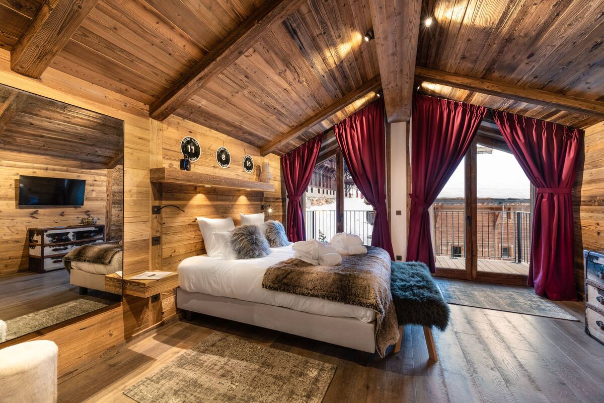 A cozy wooden cabin bedroom with a large bed, a TV, and a balcony with red curtains.