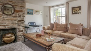 A cozy living room with a stone fireplace, a wooden coffee table with books and a candle, a sofa with decorative pillows, and a view through a large window. - Stone cottage in central Salcombe sleeps 8  (Salcombe)