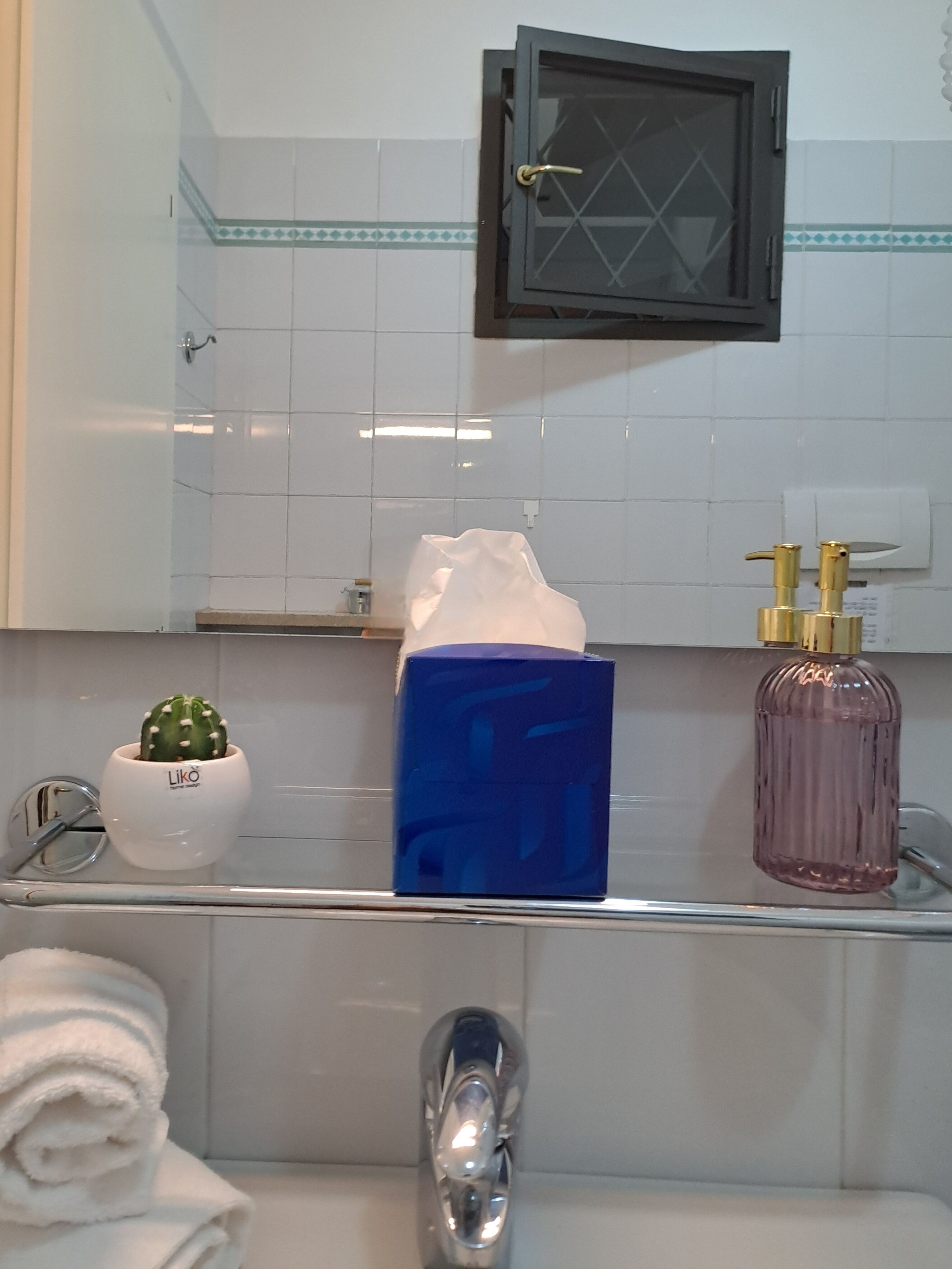 Een badkamer met een witte betegelde muur, een zwarte medicijnkast, een blauwe zakdoekendoos, een roze zeepdispenser, een kleine cactusplant en een opgerolde witte handdoek.