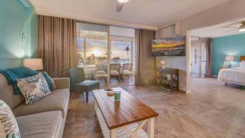 A hotel room with a sofa, a wooden table, a balcony with a view of the sunset, and a TV on the wall.