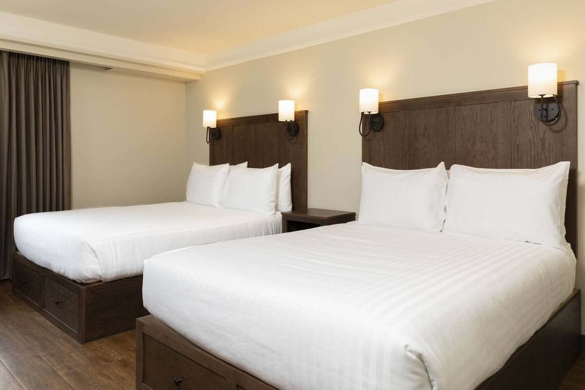 A hotel room with two single beds, wooden headboards, bedside tables, and a large window with curtains.