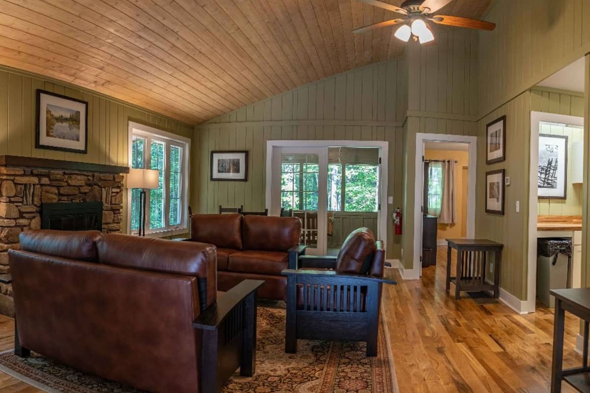 A spacious living room with a stone fireplace, leather sofas, a dark wooden coffee table, and a ceiling fan.
