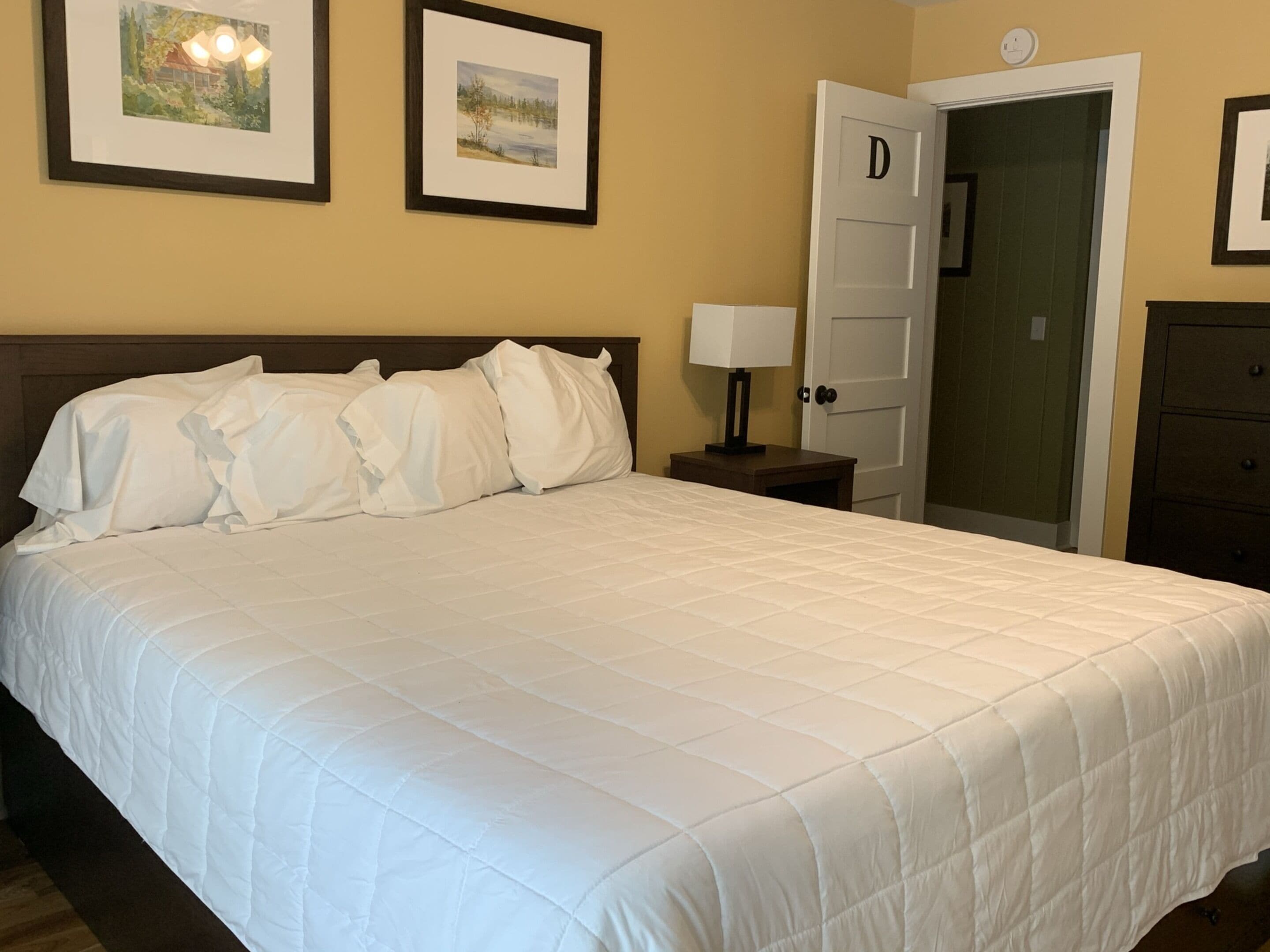Una cama bien hecha con ropa de cama blanca, una mesita de noche con una lámpara y dos cuadros en la pared.