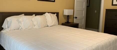 Una cama bien hecha con ropa de cama blanca, una mesita de noche con una lámpara y dos cuadros en la pared.